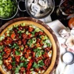 Buffalo cauliflower pizza with melted vegan mozzarella, sliced celery, and fresh cilantro, surrounded by prep ingredients on a dark table.