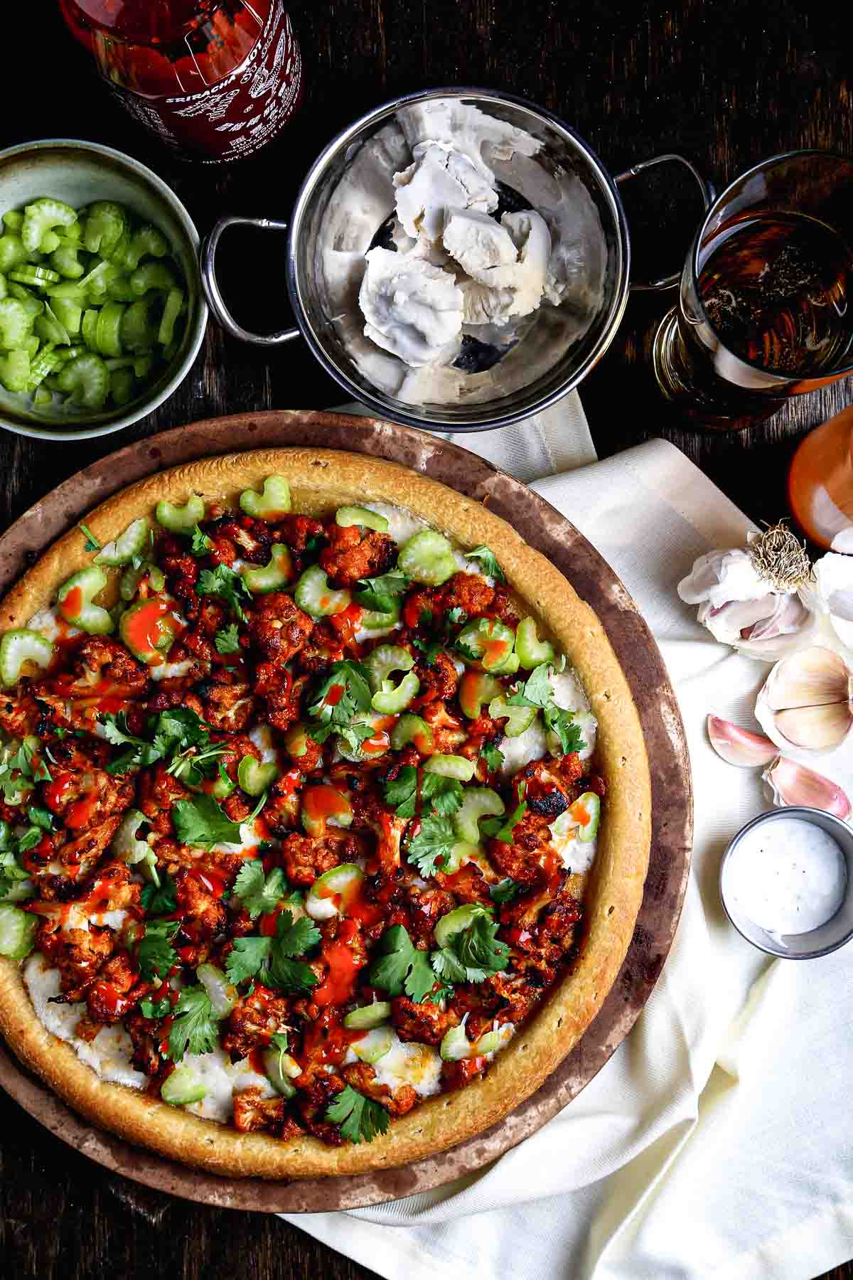 Buffalo cauliflower pizza with melted vegan mozzarella, sliced celery, and fresh cilantro, surrounded by prep ingredients on a dark table.