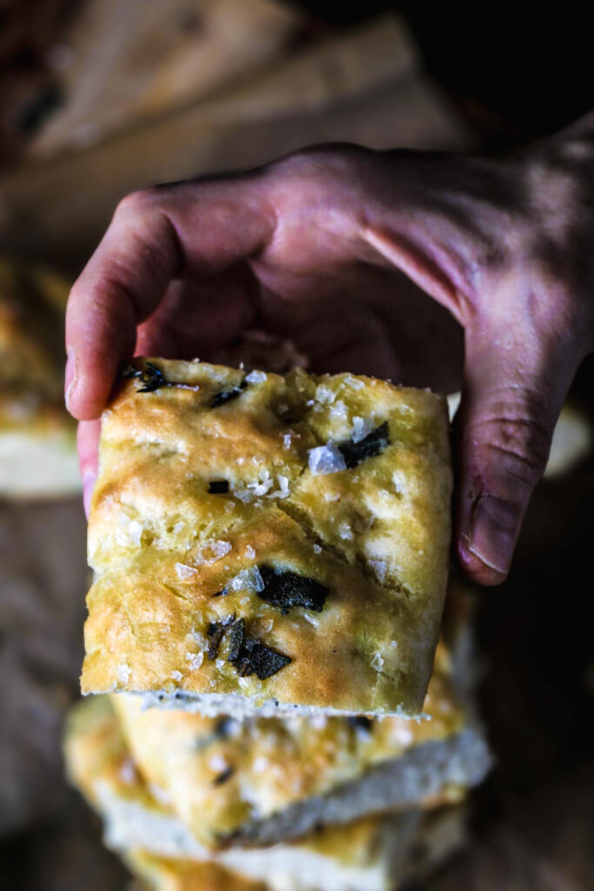 A hand holds a slice of golden-brown focaccia, topped with flaky sea salt and fresh herbs, showcasing its airy texture and crisp crust.
