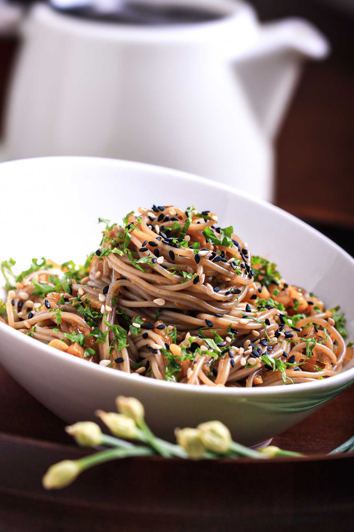 Tilted shot of an easy soba noodles recipe served in a white bowl, topped with fresh herbs and toasted sesame seeds.