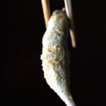 Single piece of crispy vegan fried okra held with chopsticks against a dark background.