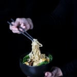 A hand holding a bowl of ramen noodles, while the other is pulling up on the noodles with chopsticks.