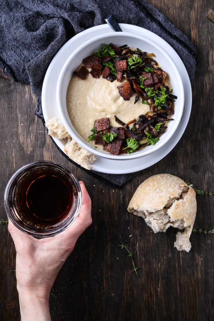 A bowl of creamy roasted parsnip soup on a wooden board. There is a piece of bread and one hand holding a glass with a dark beverage.