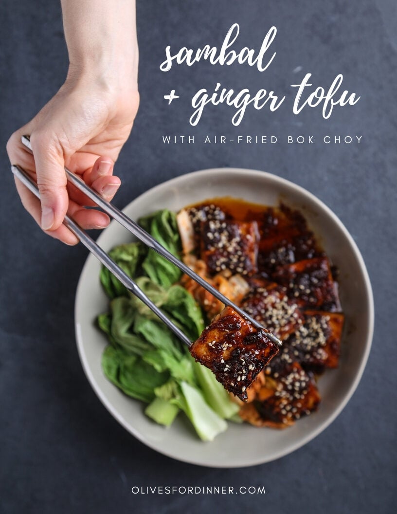 A top-down shot of spicy sambal tofu with wilted greens. One hand is holding a piece of tofu over the plate in silver chopsticks.
