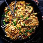 A cast iron pan with spicy peanut noodles and crispy tofu, garnished with fresh scallions, sesame seeds, and crushed peanuts, with chopsticks resting on the side.
