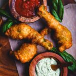 Stuffed zucchini blossoms on a wooden board with bowls of marinara and basil-cashew cheese, served hot and crispy with fresh basil garnish.