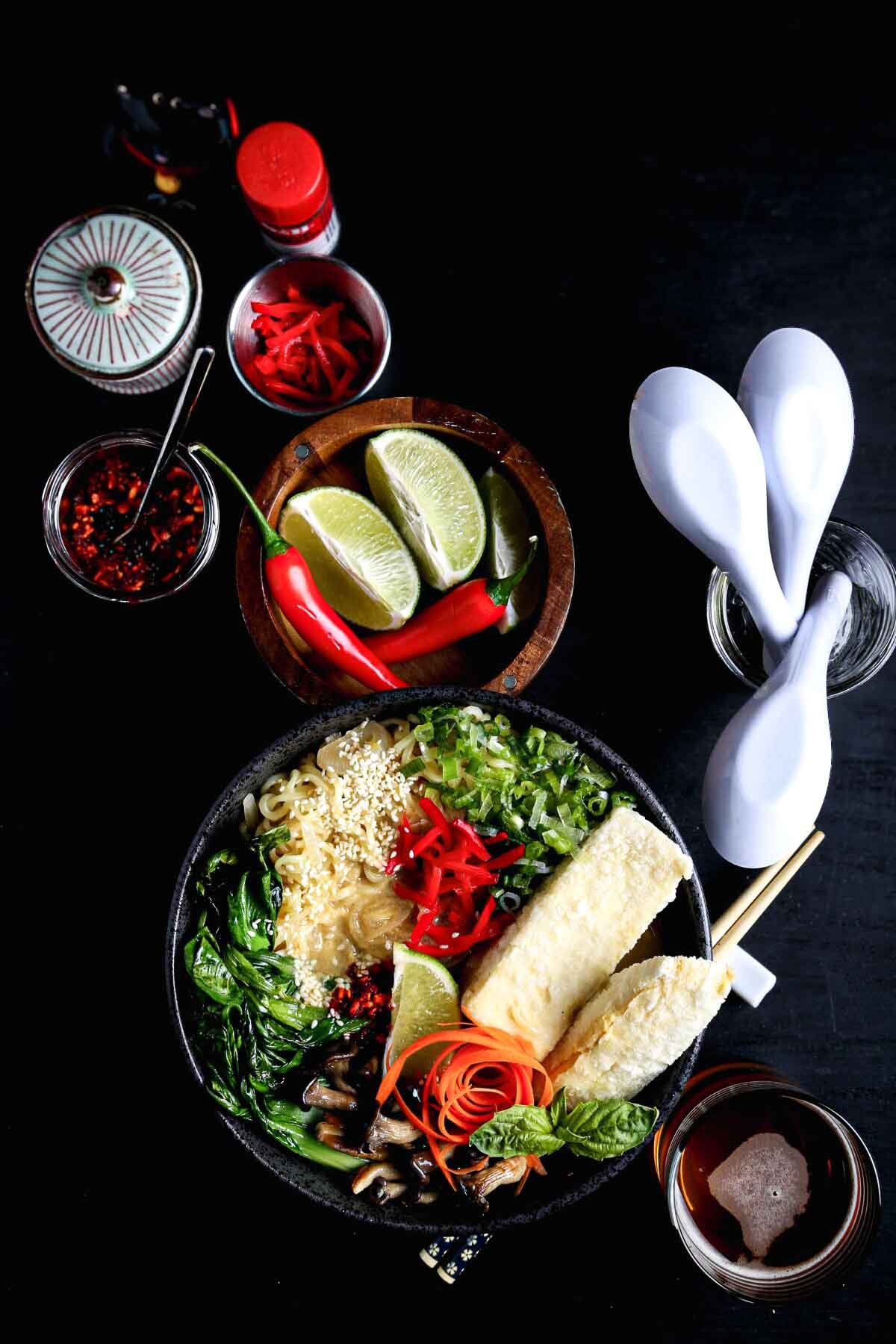 A styled vegan ramen recipe setup, featuring a bowl of ramen with toppings, chopsticks, and condiments for serving.