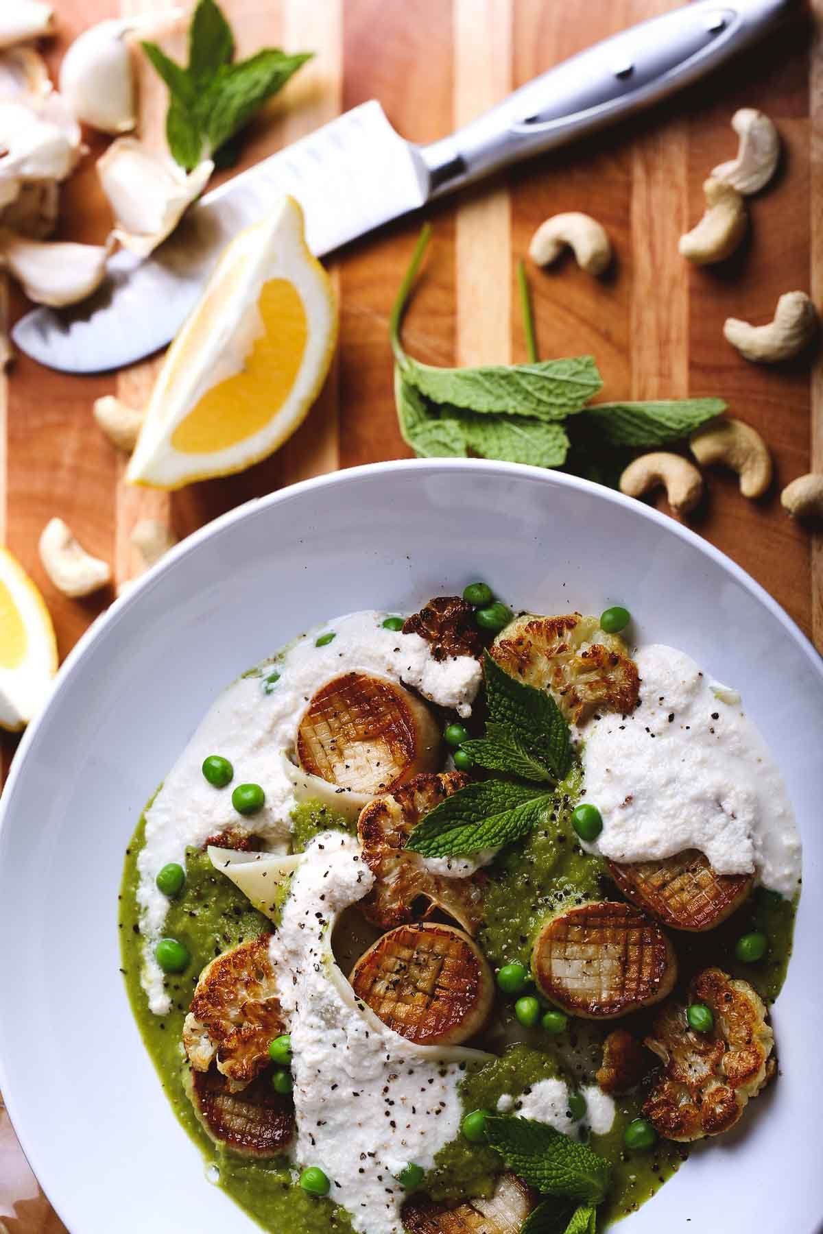 Top-down view of vegan scallops with cauliflower, mint, green pea puree, and cashew cream, surrounded by lemon, garlic, and cashews.