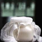 A jar of homemade vegan yogurt covered with cheesecloth, sitting on a dark surface.